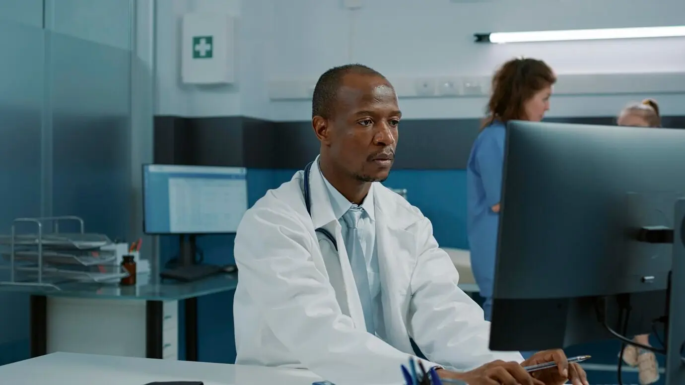 Афроамериканский врач за столом в медицинском кабинете использует компьютер для планирования приема. Медицинский специалист в белом халате и со стетоскопом занимается консультационным обслуживанием, чтобы помочь пациенту.