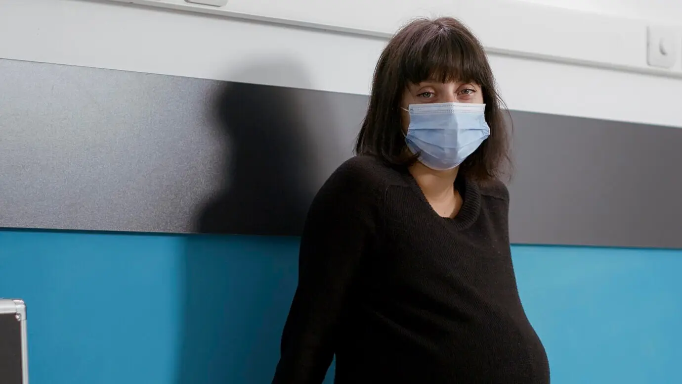 Портрет беременной пациентки в медицинском кабинете на плановом приёме у медицинского специалиста. Во время консультации женщина сидит на кушетке и в маске.